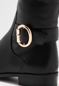 Bota de cuero negra con detalle de correa con hebilla redonda dorada y tacón bajo apilado sobre un fondo blanco.