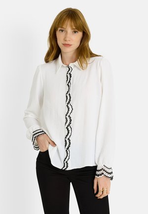 Blusa de manga larga blanca con ribete negro en forma de ondas a lo largo del frente y los puños, hecha de una tela texturizada, con un collar estándar y un corte relajado.