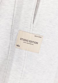 Sivá, textúrovaná tkanina s etiketou označenou "STUDIO EDITION" od Alpha Industries. Etiketa má krémové pozadie a tmavý text.