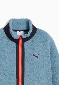 Ljusblå fleecejacka med hög krage, svarta accenter och en orange dragkedja. Har en svart Puma-logotyp på bröstet. Mjuk textur.