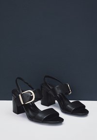 Schwarze Leder-Sandalen mit Absatz, breitem Vorderriemen, Knöchelriemen und goldener Schnallendetail. Quadratischer Blockabsatz, glatte Textur, minimalistisches Design.