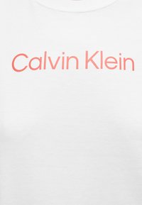 Λευκό πουκάμισο με τη φράση "Calvin Klein" γραμμένη με μεγάλα, κοραλλί γράμματα στο στήθος.