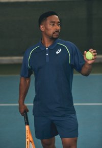 Navy sinine polo roheliste detailidega, lühikeste varrukatega ja kraega. Isik hoiab tennisepalli ja reketit väljakul.