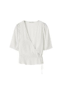 Blouse portefeuille blanche à pois noirs, manches courtes, col en V et fermeture par un lien sur le côté.