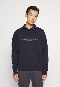 Sudadera de cuello alto con cremallera en color azul marino, con el logotipo "TOMMY HILFIGER EST. 1985" en la parte frontal. Cuenta con un bolsillo tipo canguro y puños acanalados. Material de mezcla de algodón.