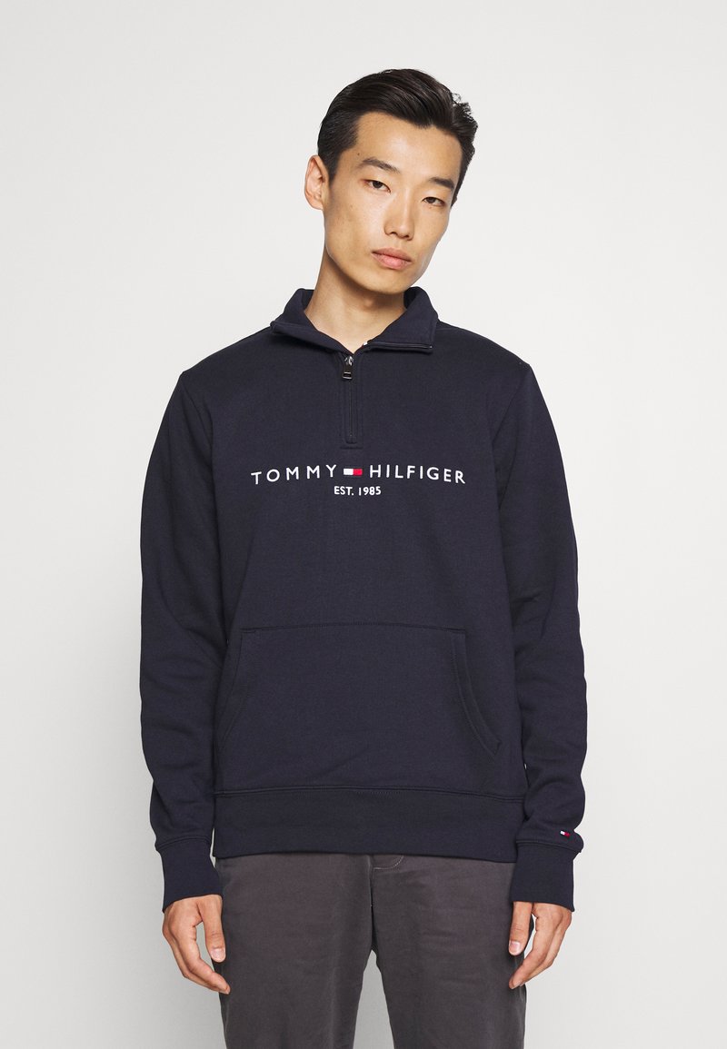 Sudadera de cuello alto con cremallera en color azul marino, con el logotipo "TOMMY HILFIGER EST. 1985" en la parte frontal. Cuenta con un bolsillo tipo canguro y puños acanalados. Material de mezcla de algodón.