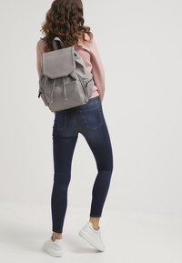 Grå läderryggsäck med snörning och framficka, bärs av en person i en rosa tröja och blå jeans, med vita sneakers.