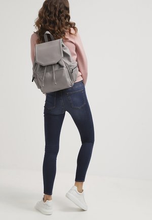 Un sac à dos en cuir gris avec une fermeture à cordon et un rabat à l'avant, porté par une personne en pull rose et en jeans bleus, avec des baskets blanches.