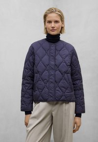 Mørkeblå quiltet jakke med rund hals, trykknaplukning og ribbede manchetter. Båret over en sort turtleneck og parret med beige bukser.