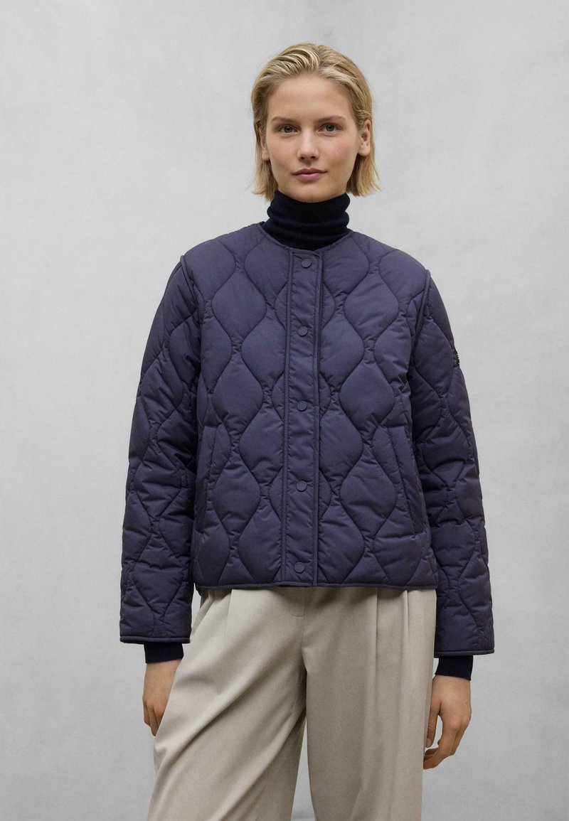 Mørkeblå quiltet jakke med rund hals, trykknaplukning og ribbede manchetter. Båret over en sort turtleneck og parret med beige bukser.