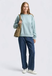 Sweat-shirt bleu clair à épaules tombantes, associé à un jean large bleu foncé. Le mannequin tient un sac marron ; des baskets blanches décontractées complètent la tenue.