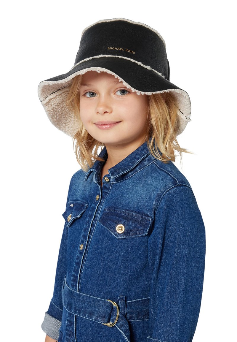 Enfant aux cheveux blonds portant un chapeau seau en shearling noir et blanc et une veste en denim bleue avec des boutons dorés et une ceinture.