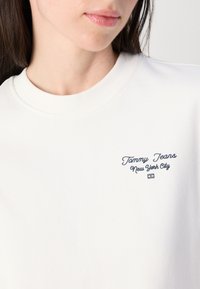 Nærbilde av en kvinnes hals og hake iført en hvithettegenser med "Tommy Jeans New York City" brodert i mørk blå tekst.
