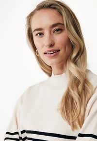 Pull en tricot crème avec un col haut, présentant des rayures horizontales bleu marine. Le mannequin a les cheveux longs et ondulés, et porte de petites boucles d'oreilles.