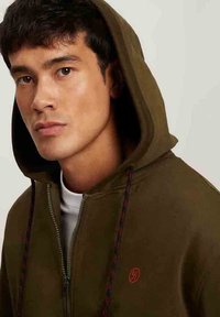 Ściągacz z zieleni oliwkowej z kapturem, zapinany na zamek, z kontrastowymi sznurkami i czerwonym logo na klatce piersiowej. Miękka struktura materiału.