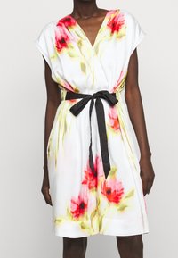 Blommönstrad klänning i vit tyg med röda och gula blommor, V-ringning, korta ärmar och ett svart bälte knutet i midjan.