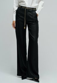 Pantalones anchos de pierna negra hechos de tela suave, con un cinturón de cadena dorada y un ajuste relajado. El diseño incluye una cintura alta y un brillo sutil.