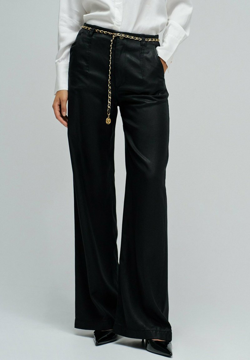 Pantalones anchos de pierna negra hechos de tela suave, con un cinturón de cadena dorada y un ajuste relajado. El diseño incluye una cintura alta y un brillo sutil.