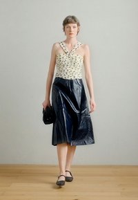 Top alb cu buline negre, cu bretele răsucite; fustă plisată, de culoare albastru închis, lucioasă; model ținând o geantă mică neagră, purtând balerini negri.