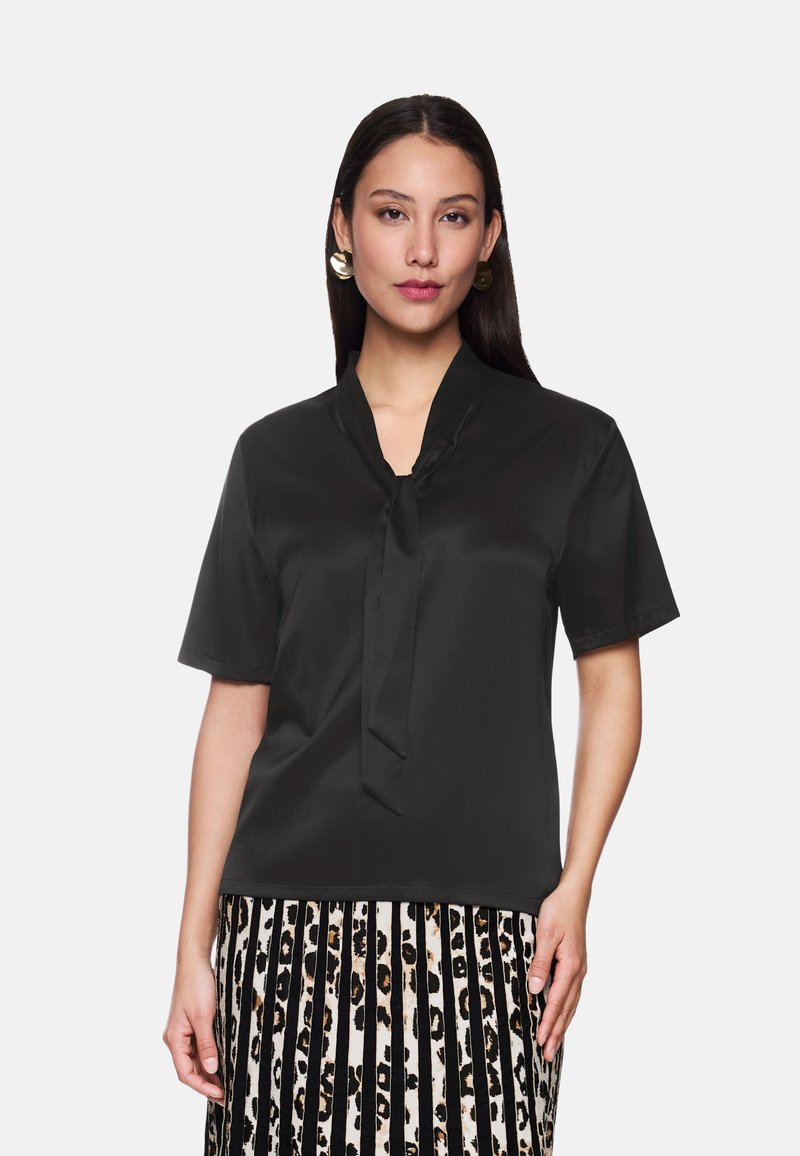 Schwarze Kurzarmbluse mit einer Schleifenkragen, aus einem glatten Stoff gefertigt. Kombiniert mit einem gestreiften Rock in Schwarz und Weiß mit einem Leopardenmuster.