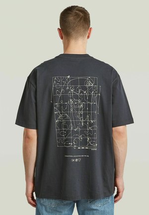 Mężczyzna zwrócony tyłem nosi ciemny t-shirt z białym abstrakcyjnym wzorem mapy i siatki na plecach, w połączeniu z niebieskimi dżinsami.