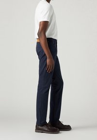 Pantaloni di cotone blu scuro con vestibilità slim, tasche laterali e una cintura in pelle marrone come dettaglio. Il modello indossa una t-shirt bianca a maniche corte.