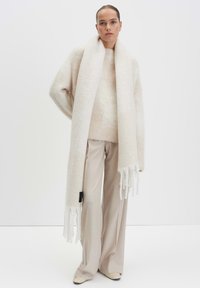 Creme wollen oversized trui met uitlopende mouwen, gecombineerd met een lange creme sjaal met franjes en bijpassende beige broek.