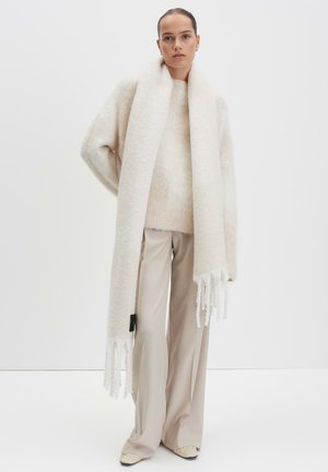 Creme wollen oversized trui met uitlopende mouwen, gecombineerd met een lange creme sjaal met franjes en bijpassende beige broek.