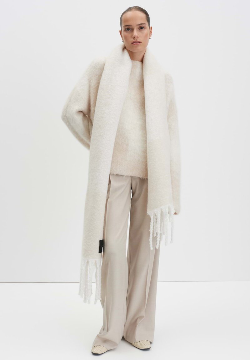 Creme wollen oversized trui met uitlopende mouwen, gecombineerd met een lange creme sjaal met franjes en bijpassende beige broek.