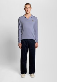 Νεαρός άνδρας που στέκεται φορώντας ένα πουλόβερ polo με μακριά μανίκια σε λεβαντίν αποχρώσεις, σκούρο μπλε παντελόνι και λευκά αθλητικά παπούτσια μπροστά σε ένα απλό φωτεινό φόντο.