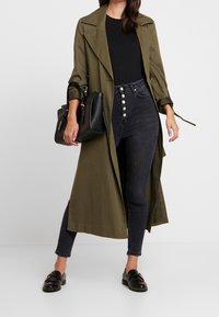Abrigo trench de color verde oliva con cuello y apertura frontal, combinado con jeans negros de cintura alta con detalles de botones y mocasines negros.