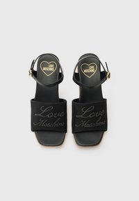 Μαύρες σαγιονάρες με χοντρό τακούνι, με μια φαρδιά υφασμάτινη λωρίδα διακοσμημένη με το "Love Moschino" σε καλλιγραφία, και μια μικρή λωρίδα στον αστράγαλο με αγκράφα.