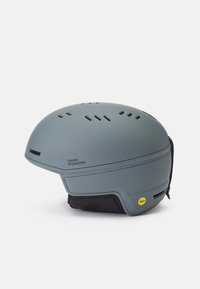Un casco de esquí gris presenta un diseño suave y redondeado con agujeros de ventilación, correas ajustables y una etiqueta que indica tecnología MIPS.