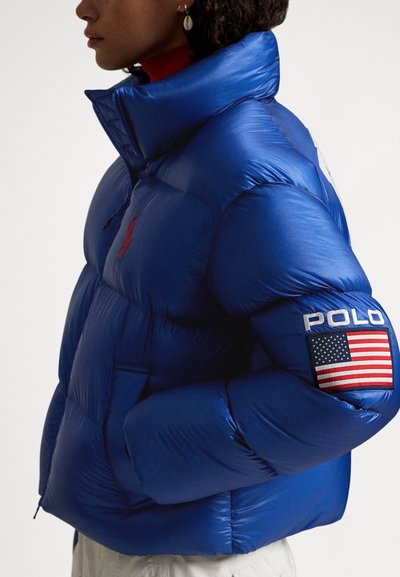 Blue puffer jacket featuring a high collar, with a red emblem on the chest and a patch on the sleeve displaying "POLO" and the American flag.
