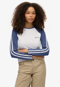 Koszulka z rękawami raglan w kolorze białym i granatowym z niebieskimi paskami, z logo "Superdry" na piersi, zestawiona z beżowymi spodniami.