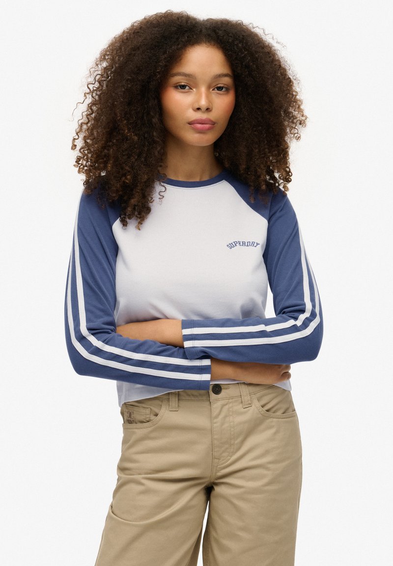 Koszulka z rękawami raglan w kolorze białym i granatowym z niebieskimi paskami, z logo "Superdry" na piersi, zestawiona z beżowymi spodniami.