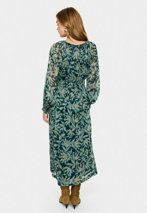 Eine von hinten fotografierte Frau trägt ein grün-blumiges Midi-Kleid mit langen Ärmeln und Smocking-Taille sowie braune Wildlederstiefel vor einem weißen Hintergrund.