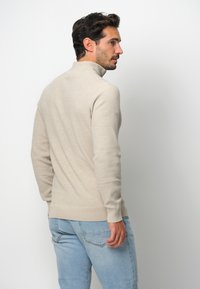 Beige, texturerad turtlenecktröja med en åtsittande design, ribbade manschetter och nederkant, matchad med ljusblå jeans, sedd bakifrån.