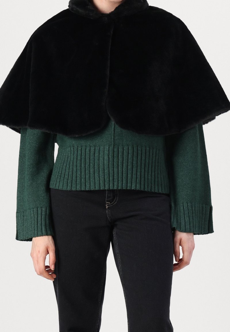 Personne portant une cape en fourrure noire sur un pull vert côtelé et un pantalon noir, montrée du cou jusqu’aux hanches sur un fond uni.
