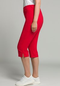Pantalons rouges courts en matériau extensible, avec un ourlet fendu. Portés avec des chaussures de sport blanches, présentés de côté.