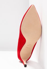 Chaussure à talon haut en daim rouge à bout pointu vue de la semelle avec un talon kitten fin reposant contre une surface blanche en angle.