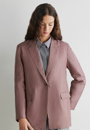 Kobieta z długimi falującymi włosami, ubrana w mauve marynarkę na jeden guzik na szarej koszuli z kołnierzykiem, patrząca w dół na jednolitym białym tle.