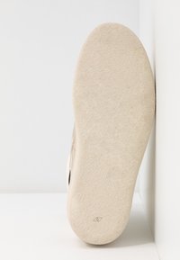 Semelle d'une chaussure en daim beige sur fond blanc, montrant une texture lisse et un petit logo embossé près du talon.