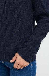 Sötétkék kötött pulóver, texturált felülettel és széles mandzsettákkal. Farmernadrággal párosítva, hangsúlyozva a laza szabást és a puha anyagot.