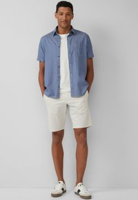 Jasnoniebieska koszula z krótkim rękawem i guzikami, z kieszonką, noszona na białej koszulce, w zestawie z beżowymi szortami i białymi trampkami.