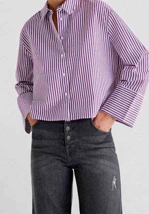 Kvinde iført en lilla og hvid verticalstribet skjorte med knapper og lange ærmer samt sorte højtaljede jeans med knapper og en lille rift.