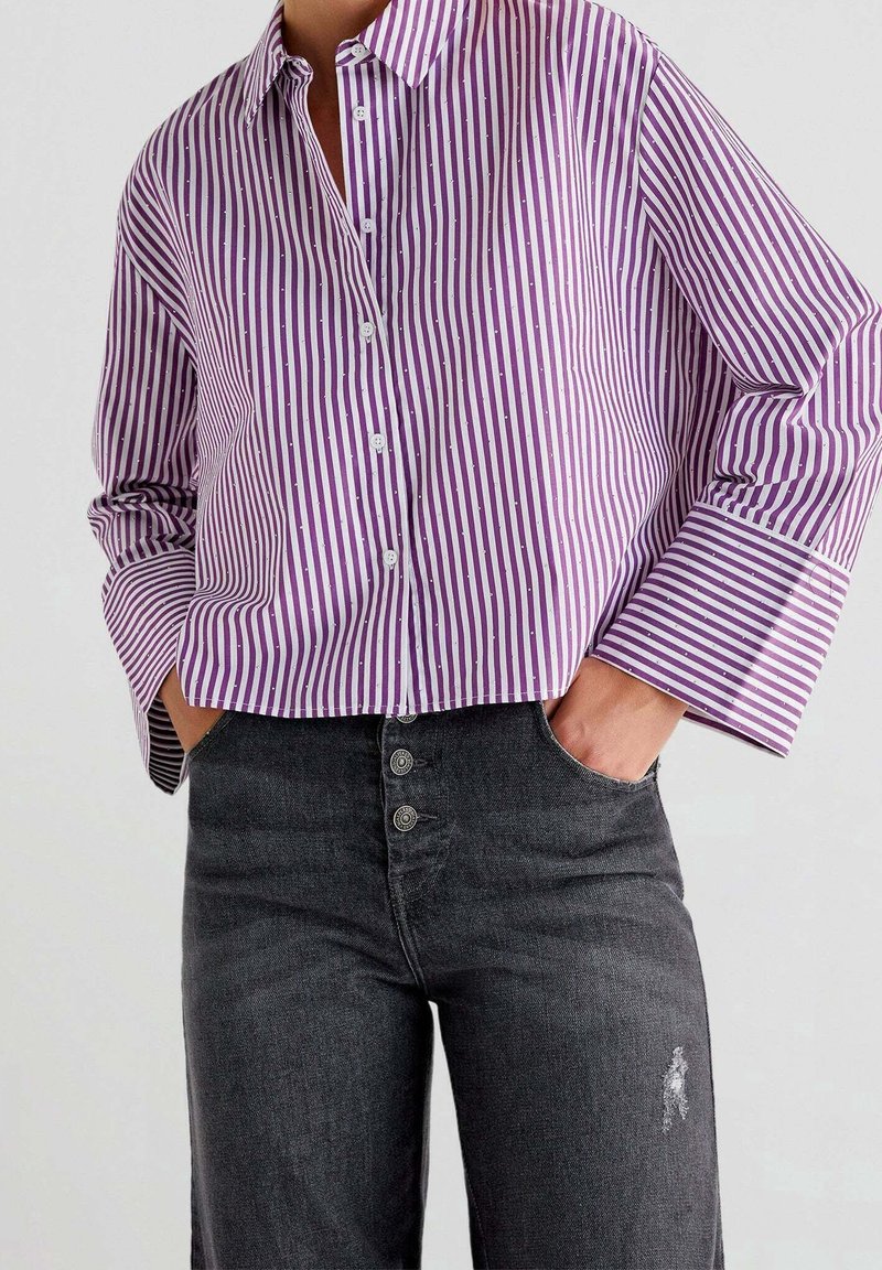 Femme portant une chemise à boutons à rayures verticales violettes et blanches, à manches longues, et un jean noir taille haute avec des boutons et une petite déchirure.