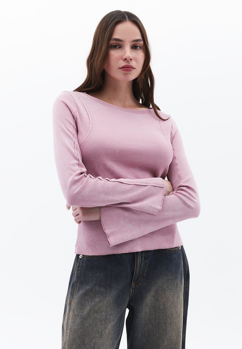 Helles rosa Langarmshirt mit rundem Ausschnitt, das subtile Nahtdetails und ausgestellte Ärmel hat, kombiniert mit dunkelblauen Jeans.