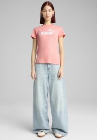 Rožinė trumpomis rankovėmis marškinėliai su balta "PUMA" logotipu, derinami su šviesiai mėlynomis plačiomis kelnėmis ir baltomis bateliais su rudais padais.