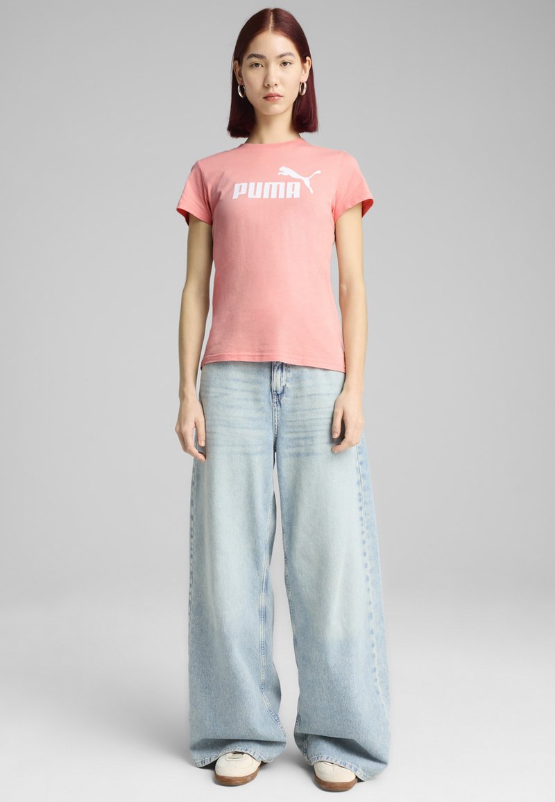 Rožinė trumpomis rankovėmis marškinėliai su balta "PUMA" logotipu, derinami su šviesiai mėlynomis plačiomis kelnėmis ir baltomis bateliais su rudais padais.